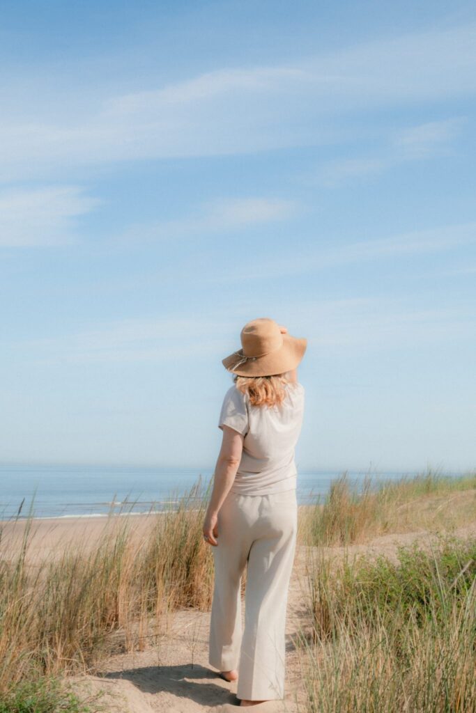 Barefoot woman walking away along a sandy dune path, holding a sun hat beneath a clear blue sky. Noordwijk - Contemporary artist Leonoor Ruigrok